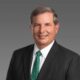 Professional headshot middle aged man in a dark suit and white shirt with a green patterned tie smiling against a gray gradient background