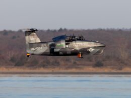 Military tilt rotor aircraft in gray camouflage flying low over a calm body of water with a forested shore in the background markings read SQU 0134 and REGENT DEFENSE