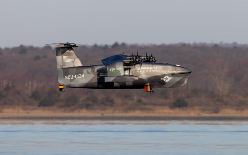 69dce7216842738c85a5048a IMG 6517HQ Military tilt rotor aircraft in gray camouflage flying low over a calm body of water with a forested shore in the background markings read SQU 0134 and REGENT DEFENSE