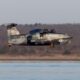 Military tilt rotor aircraft in gray camouflage flying low over a calm body of water with a forested shore in the background markings read SQU 0134 and REGENT DEFENSE
