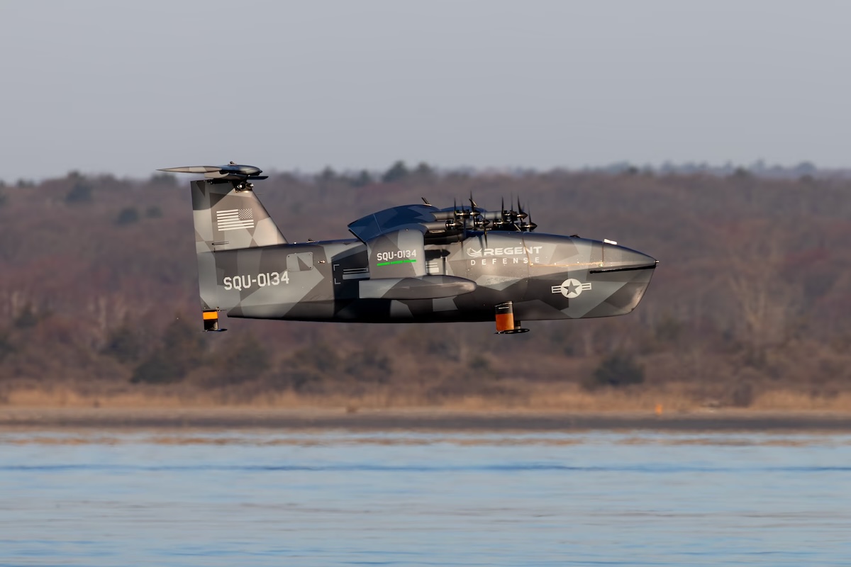 Military tilt rotor aircraft in gray camouflage flying low over a calm body of water with a forested shore in the background markings read SQU 0134 and REGENT DEFENSE