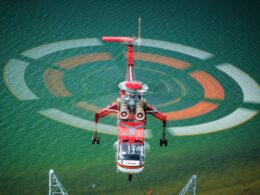 Red rescue helicopter hovering over turquoise water rotor blur forming concentric circles above the sea with a hoist line visible