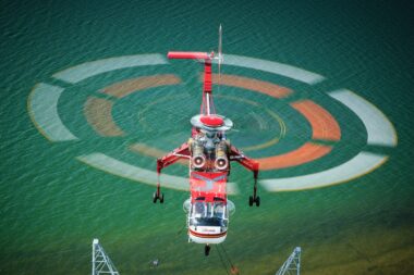 Red rescue helicopter hovering over turquoise water rotor blur forming concentric circles above the sea with a hoist line visible