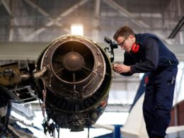 Engineer in blue overalls inspecting a jet engine in a hangar wearing safety goggles and red ear protection