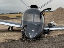 Small grey airplane crashed on a gravel surface nose damaged and cockpit canopy intact wings wide and landing gear damaged nearby