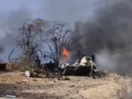 Burning wreckage on a dry barren ground with blackened trees and thick smoke