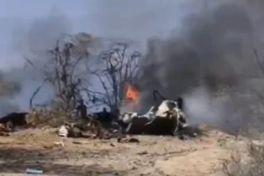 Burning wreckage on a dry barren ground with blackened trees and thick smoke