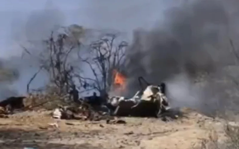 Burning wreckage on a dry barren ground with blackened trees and thick smoke