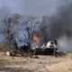Burning wreckage on a dry barren ground with blackened trees and thick smoke