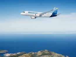 White passenger jet with a blue discover logo flying over a calm blue sea and rocky coastline below