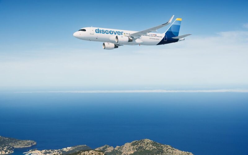 Discover A320 200 White passenger jet with a blue discover logo flying over a calm blue sea and rocky coastline below