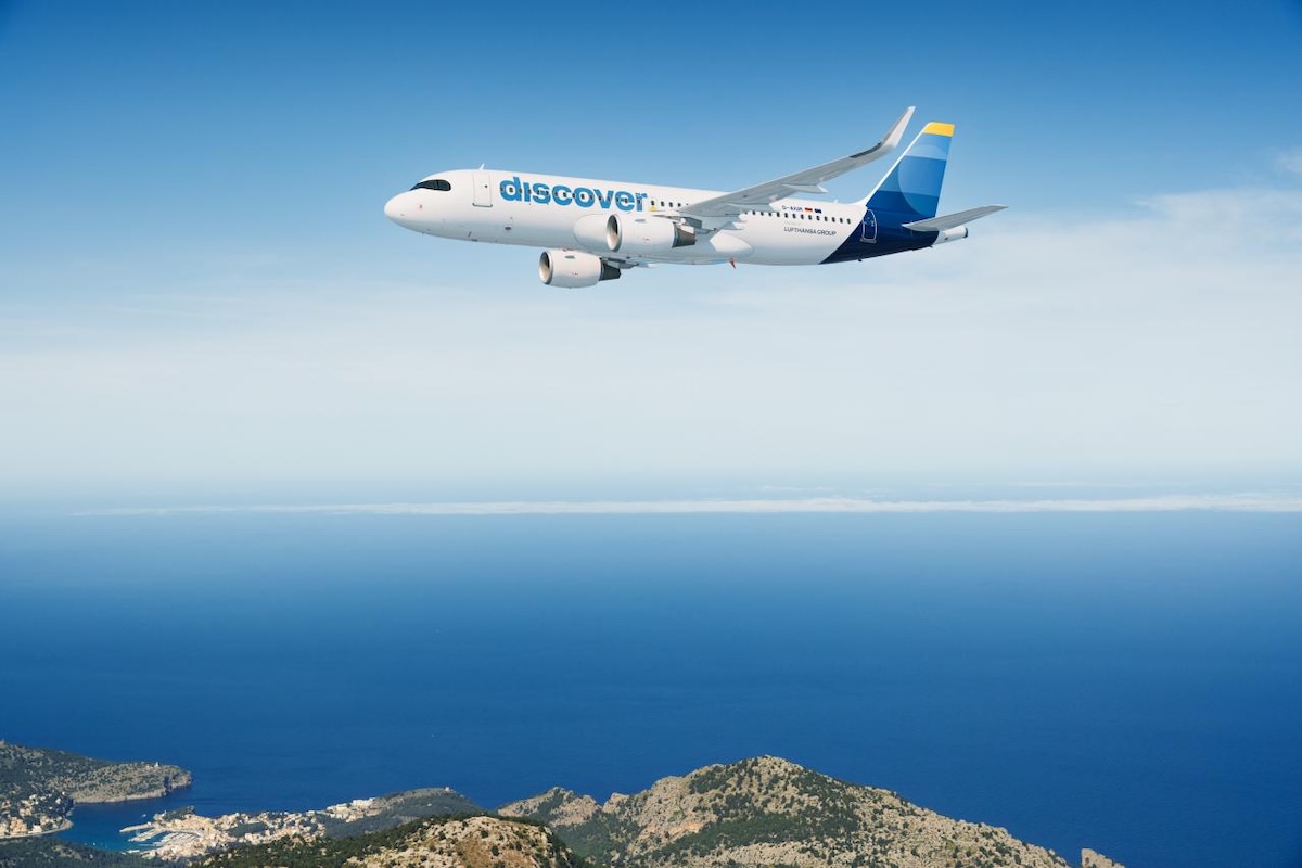 Discover A320 200 White passenger jet with a blue discover logo flying over a calm blue sea and rocky coastline below