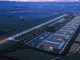 Aerial view of a large industrial campus at dusk with long warehouse buildings solar panels on roofs and a central illuminated access road or runway lined with lights