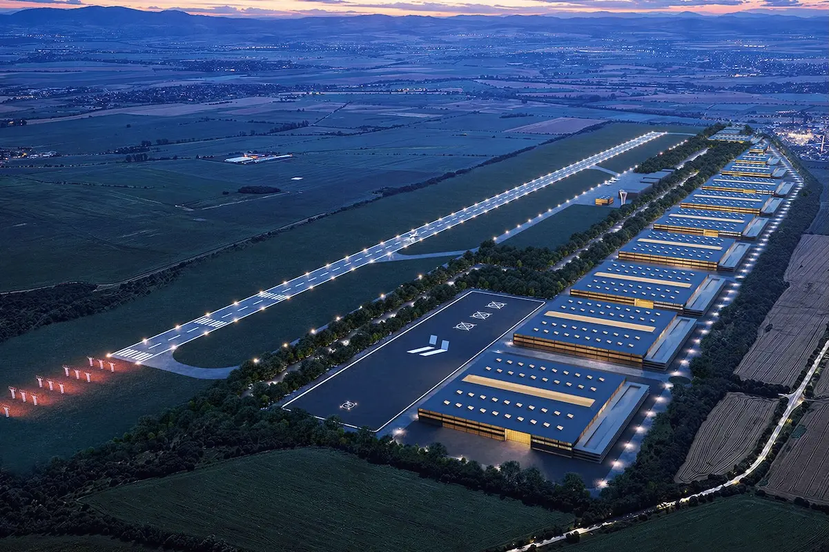 Aerial view of a large industrial campus at dusk with long warehouse buildings solar panels on roofs and a central illuminated access road or runway lined with lights