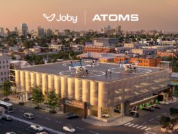 Aerial view of a modern Mobility Hub building with two rooftop helicopters and a city skyline showing Joby and ATOMS branding