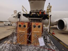 Open cargo hold of a passenger jet being loaded with large wooden crates secured by nets workers in orange vests stand on the platform above the crates on a loading rig