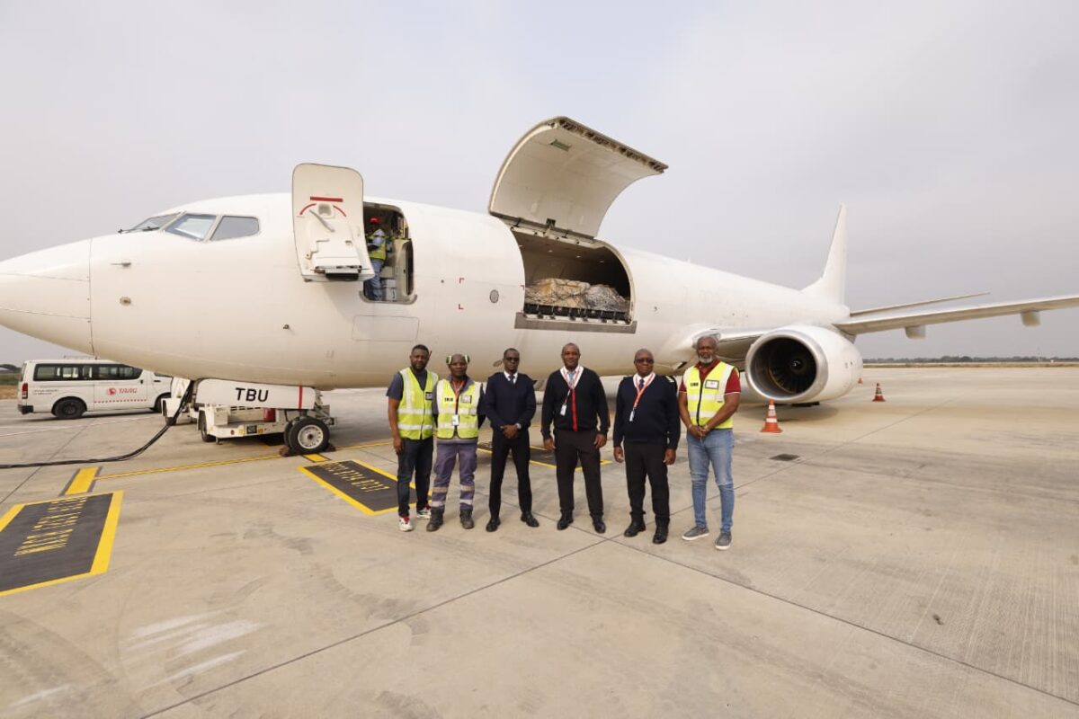 Group of airline ground crew and pilots standing on runway beside a white cargo plane with an open aft cargo door on the tarmac