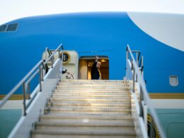 Man in a dark suit waves from the open doorway of a blue airplane at the top of metal stairs