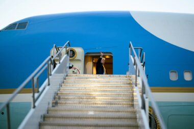 Man in a dark suit waves from the open doorway of a blue airplane at the top of metal stairs