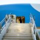 Man in a dark suit waves from the open doorway of a blue airplane at the top of metal stairs