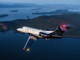 Private jet in flight over a blue sea with forested islands and distant mountains at dusk tail number N373EE visible on the fuselage