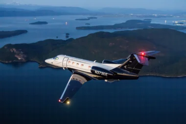 Private jet in flight over a blue sea with forested islands and distant mountains at dusk tail number N373EE visible on the fuselage