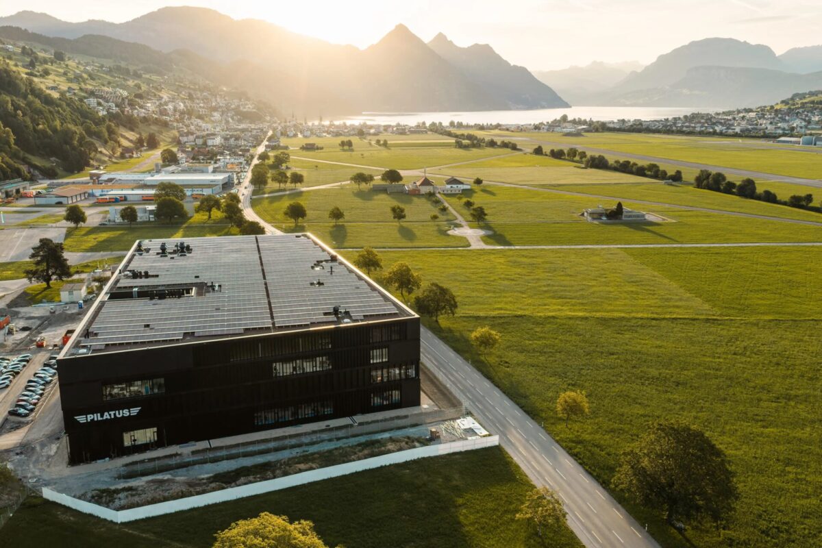 Aerial view of a modern black building with a large solar panel roof surrounded by green fields and distant mountains by a lake