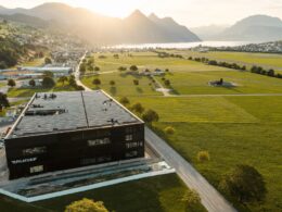 Aerial view of a modern black building with a large solar panel roof surrounded by green fields and distant mountains by a lake