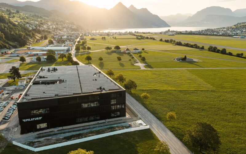 Schwarzhorn 1 Aerial view of a modern black building with a large solar panel roof surrounded by green fields and distant mountains by a lake
