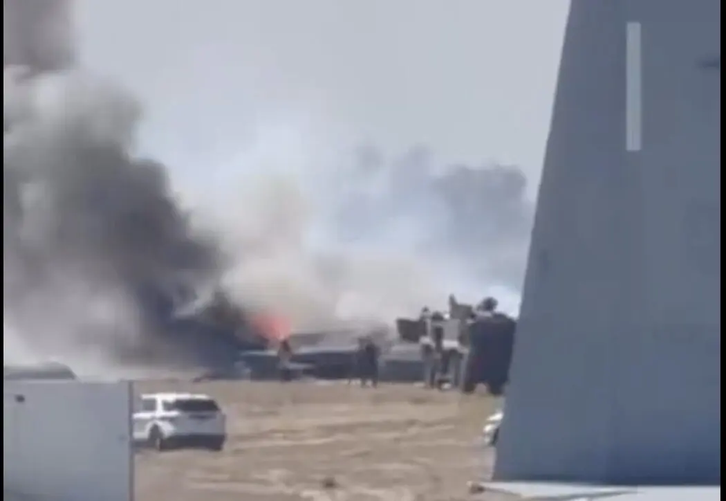 Screenshot 2026 08 04 at 20116 PM Explosion with dark smoke rising from a wreck on a dusty field several people and responders nearby