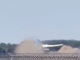 Dust and smoke plume rising from a blast behind a chain link fence near a tree line