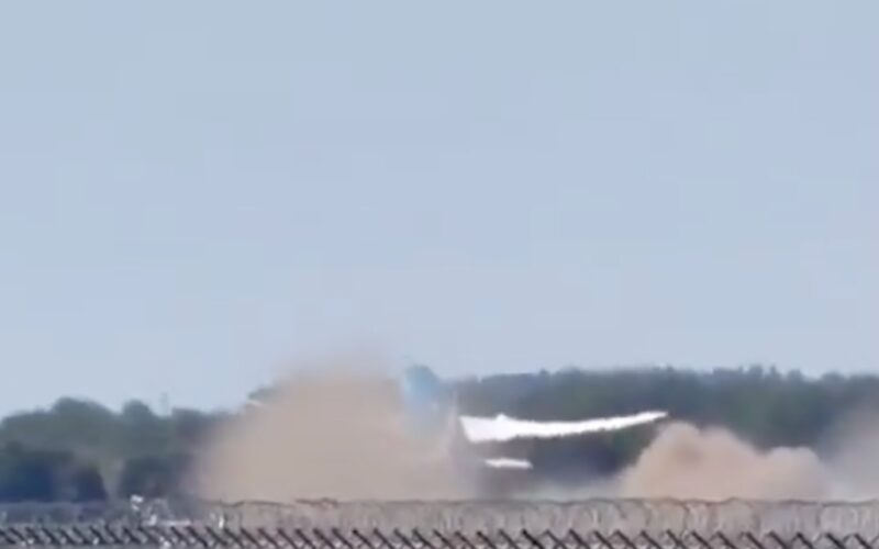Screenshot Dust and smoke plume rising from a blast behind a chain link fence near a tree line