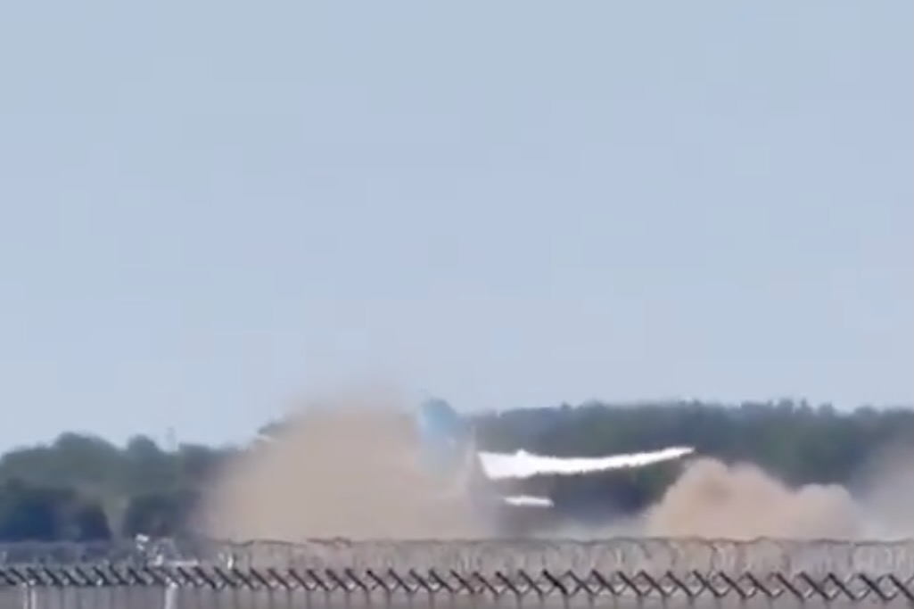 Dust and smoke plume rising from a blast behind a chain link fence near a tree line