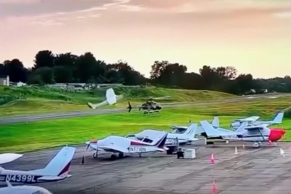Several small airplanes parked on a runway at an airfield with a helicopter on the grass and a person in white nearby during sunset