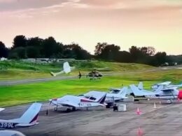 Several small airplanes parked on a runway at an airfield with a helicopter on the grass and a person in white nearby during sunset