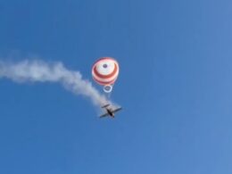 Skydiver under a red and white round parachute with a smoke trail behind a small airplane in a clear blue sky