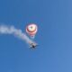 Skydiver under a red and white round parachute with a smoke trail behind a small airplane in a clear blue sky