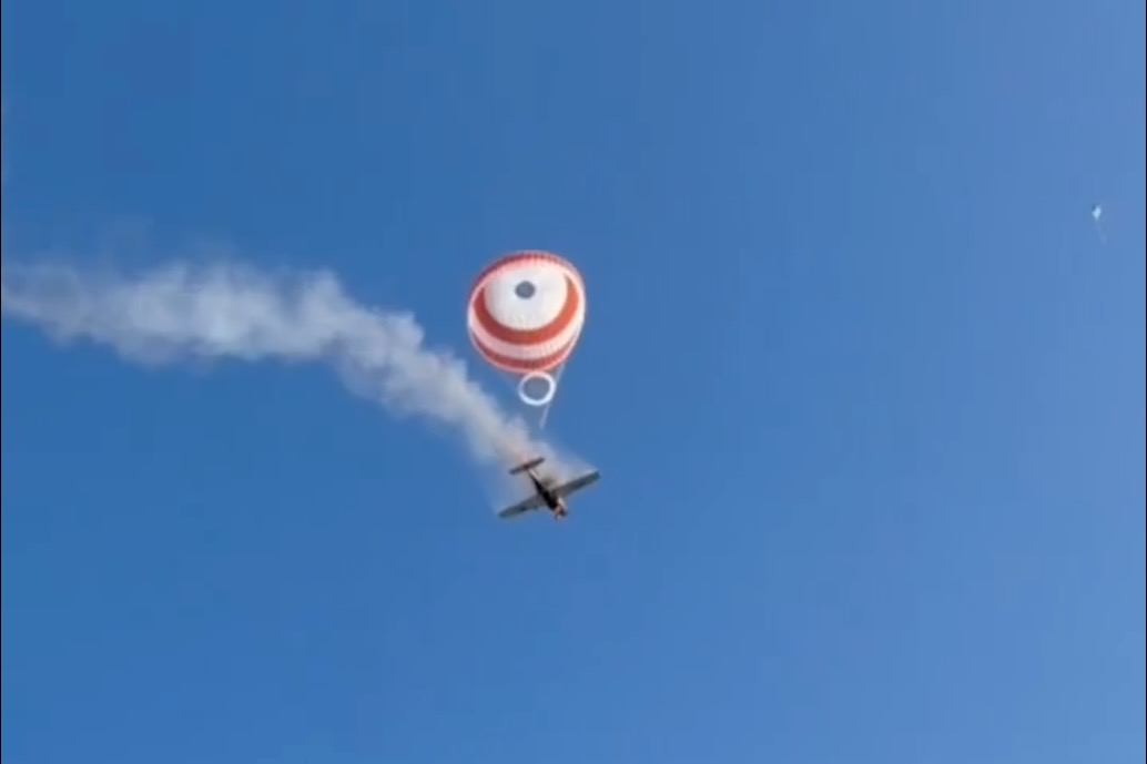 Skydiver under a red and white round parachute with a smoke trail behind a small airplane in a clear blue sky