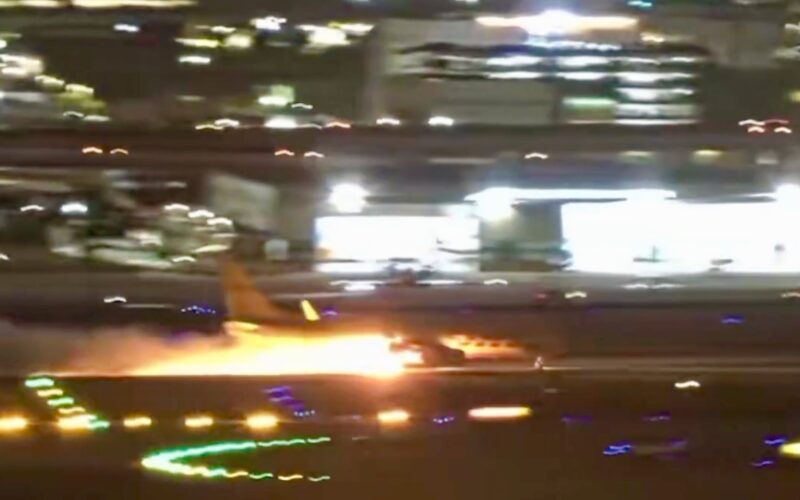 Screenshot Nighttime airport scene a blurred aircraft on the runway with bright flames or exhaust nearby and city lights in the background on a hazy night
