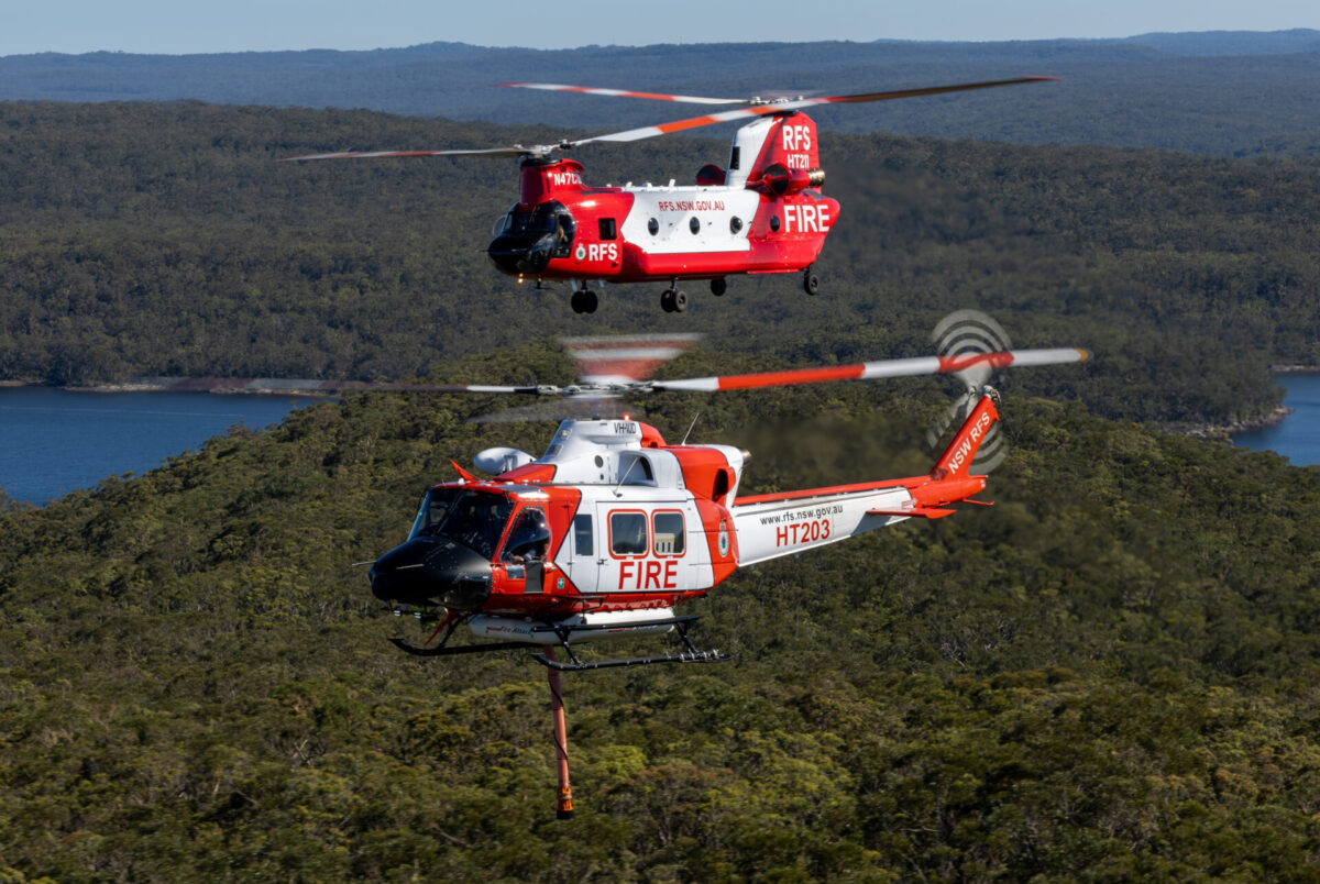 The NSW Rural Fire Services Bell 412 HeliTak 203 VH VJD foreground and CH 47D Chinook HeliTak 211 N47CU fly in formation Two redandwhite firefighting helicopters hover above a forested landscape a rope hoist descending toward the trees