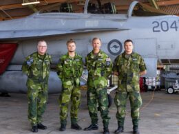 Four air force personnel in camouflage uniforms stand in front of a fighter jet inside a hangar