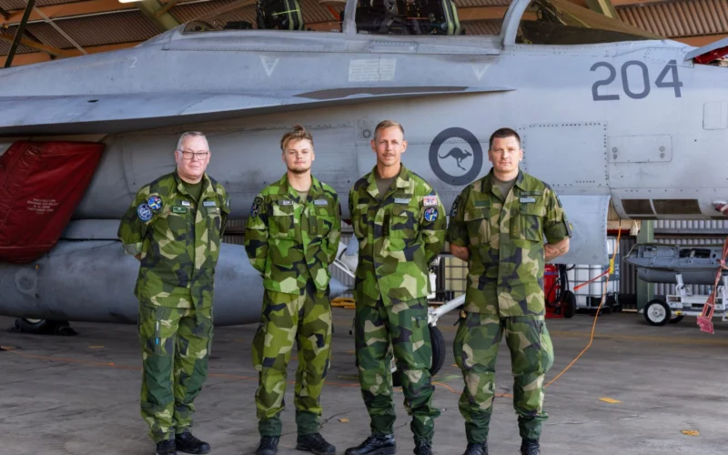 Four air force personnel in camouflage uniforms stand in front of a fighter jet inside a hangar