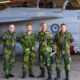 Four air force personnel in camouflage uniforms stand in front of a fighter jet inside a hangar