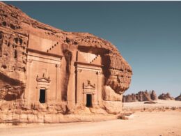 Ancient sandstone temple carved into a red cliff featuring arched doorways and decorative pediments in a desert landscape