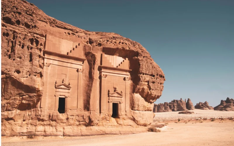 Ancient sandstone temple carved into a red cliff featuring arched doorways and decorative pediments in a desert landscape
