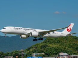 Japan Airlines passenger jet with landing gear down flying low above green hills against a blue sky