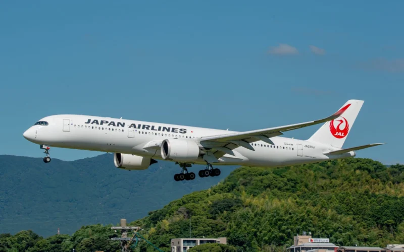 Japan Airlines passenger jet with landing gear down flying low above green hills against a blue sky