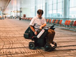 Man in a light shirt sits on a mobility scooter in an airport terminal looking at the control paneltablet in his hands