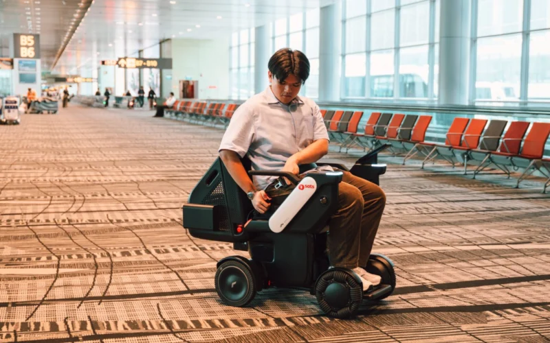 Man in a light shirt sits on a mobility scooter in an airport terminal looking at the control paneltablet in his hands