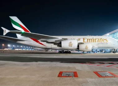 Emirates wide-body jet parked at night on an airport tarmac with terminal building in the background.